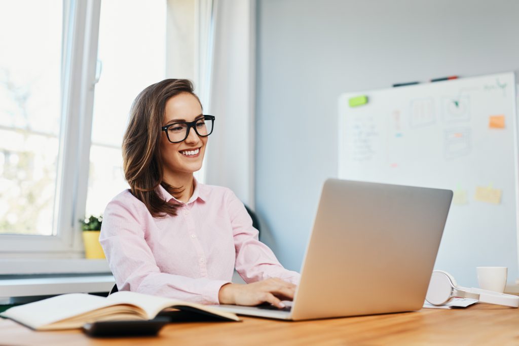 Mujer con lentes y pelo largo, escribiendo en una computadora y sentada en un escritorio.