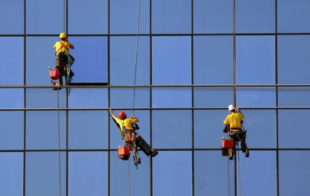 Imagen de tres personas que están colgadas de un edificio para limpiar las ventanas del mismo, están vestidos de amarillo con cascos de diferentes colores.