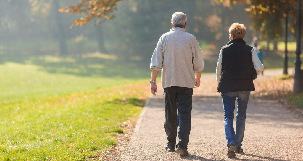 Una abuela y un abuelo caminando juntos por el parque donde hay arboles y pasto.